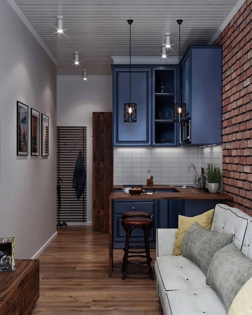 Small studio apartment interior with blue kitchen cabinets, white sofa, and brick wall.