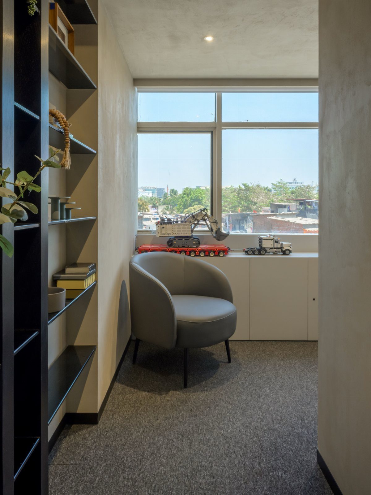 Small office nook with a grey armchair, black bookshelf, and large city window.