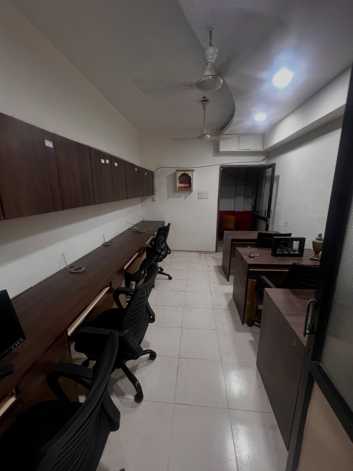 Narrow office room with brown wooden desks, black chairs, and white tiled floors.