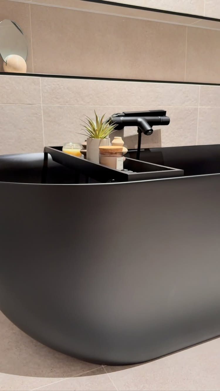 Matte black freestanding bathtub with a black caddy against beige tiled walls