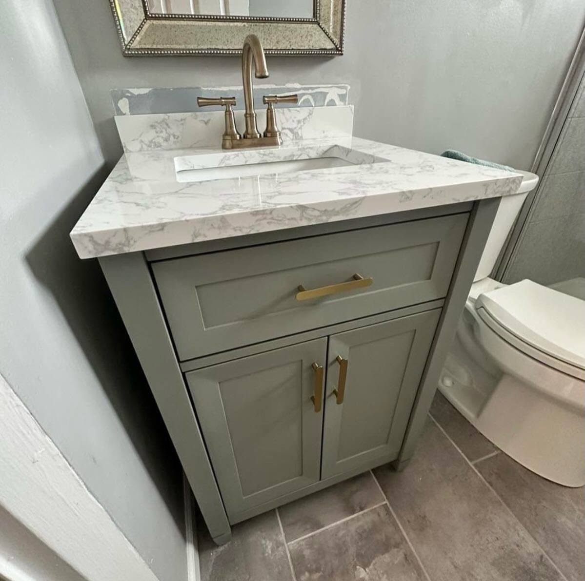 Grey freestanding bathroom vanity with white marble countertop and gold hardware next to toilet