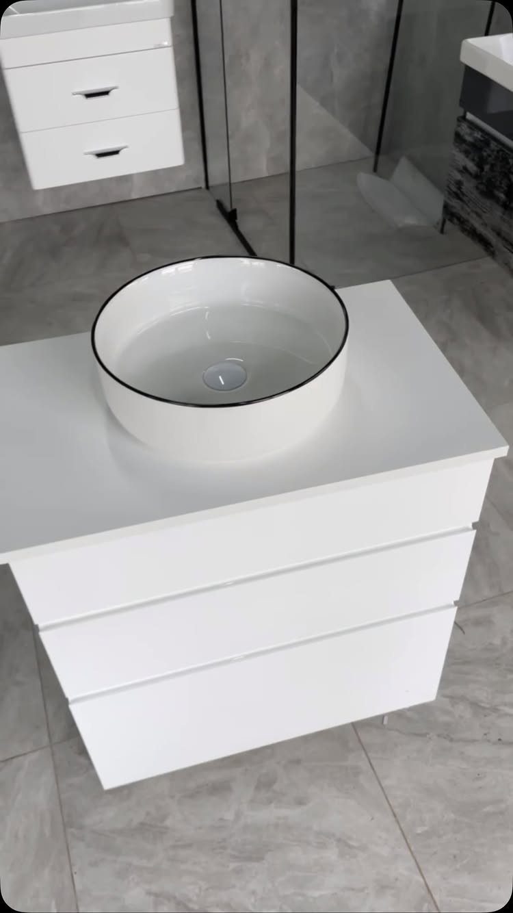 White bathroom vanity with three drawers and a black-rimmed vessel sink on marble tiles.