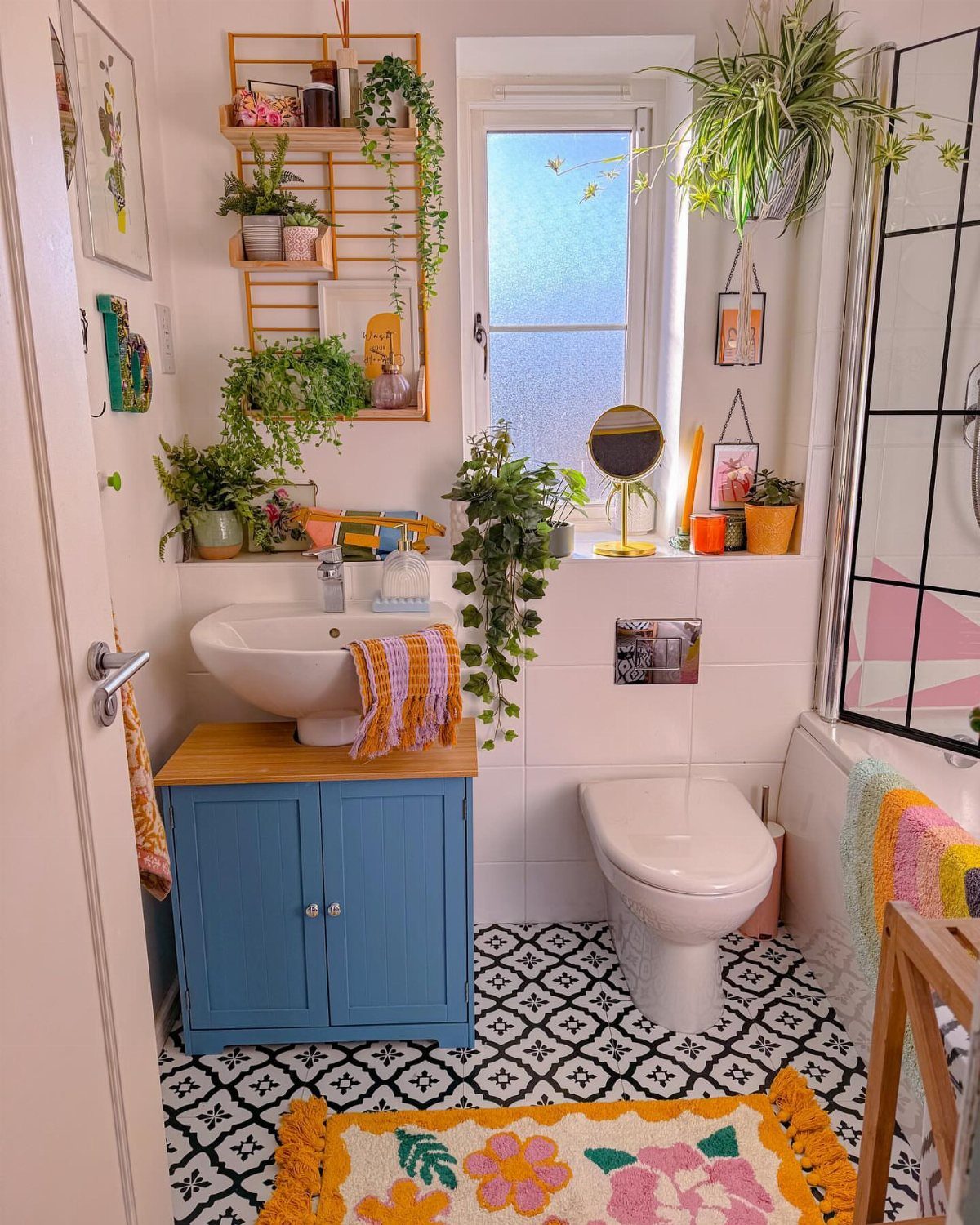 Small bathroom with blue vanity, white toilet, patterned floor, and many indoor plants.