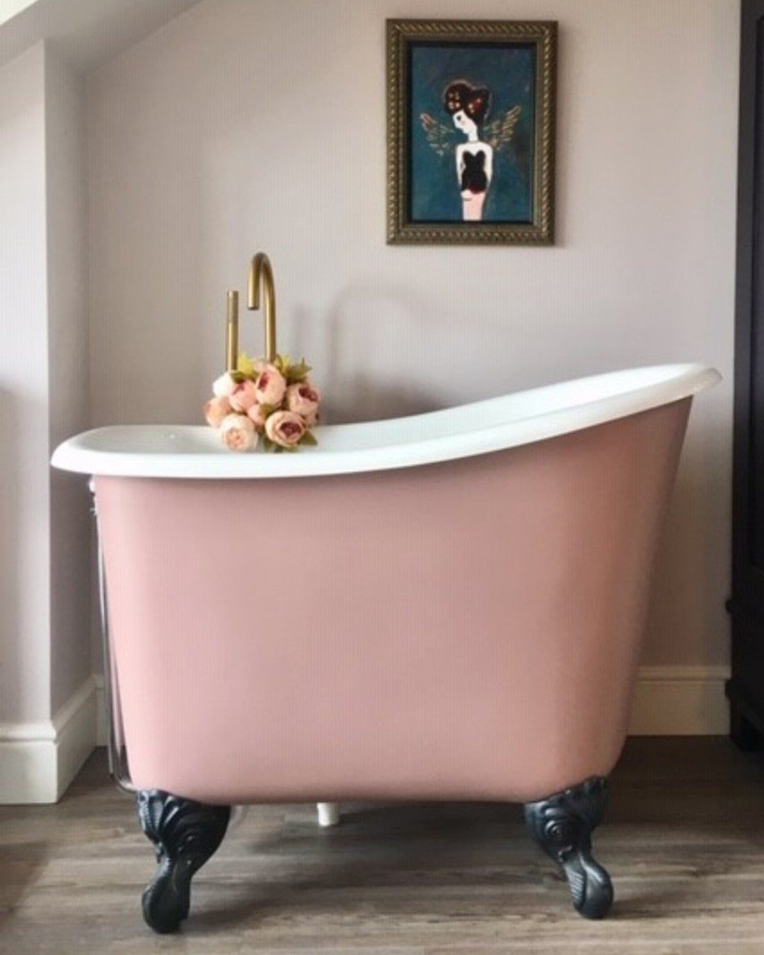 Pink clawfoot freestanding bathtub with gold faucet and roses in a bright room.