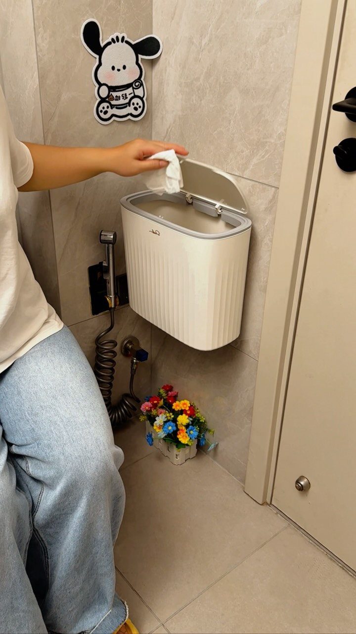 White wall-mounted trash bin in a small bathroom with beige tiles and flowers.