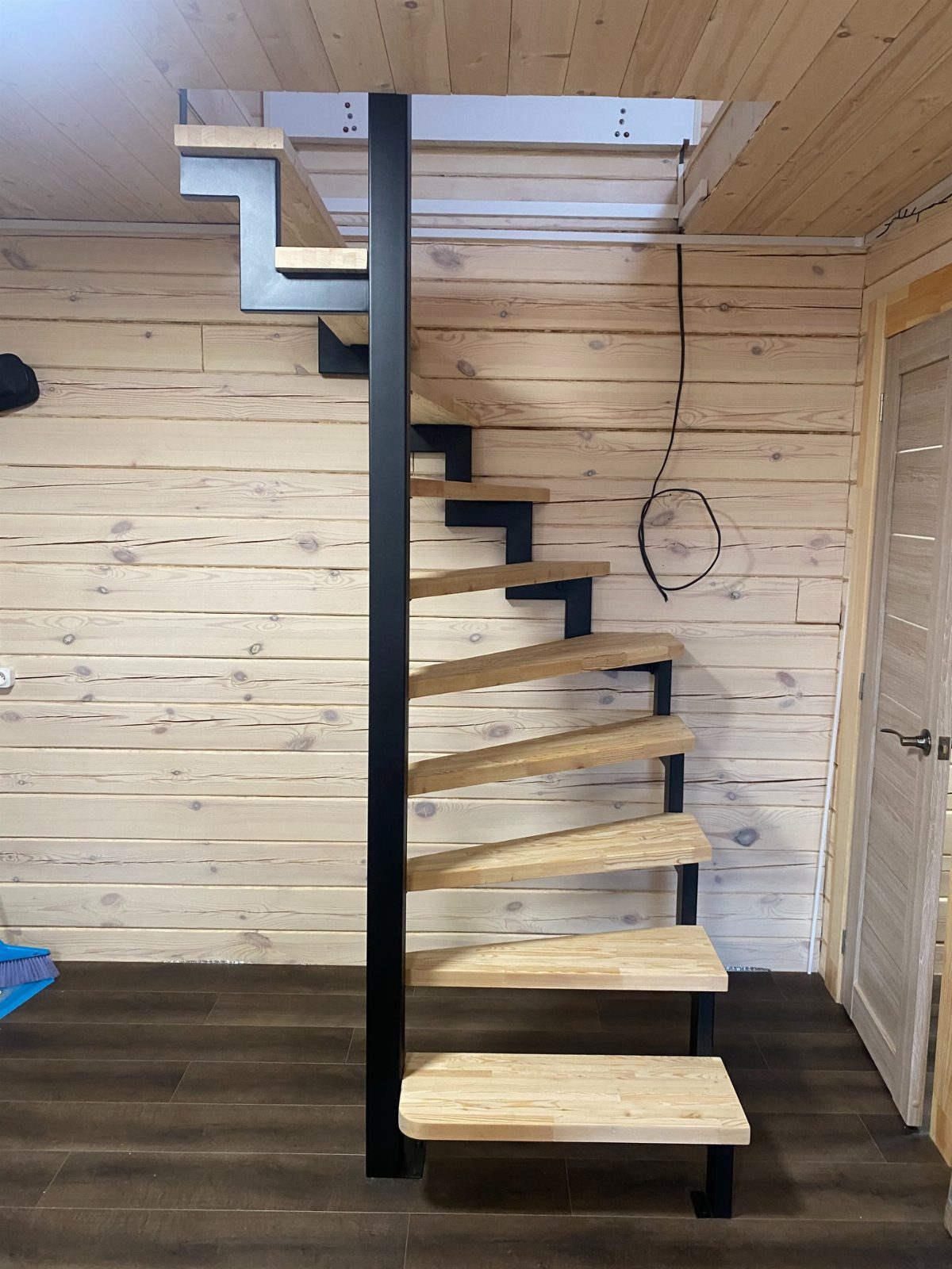 Spiral staircase with wooden steps and black metal frame against light wood walls.