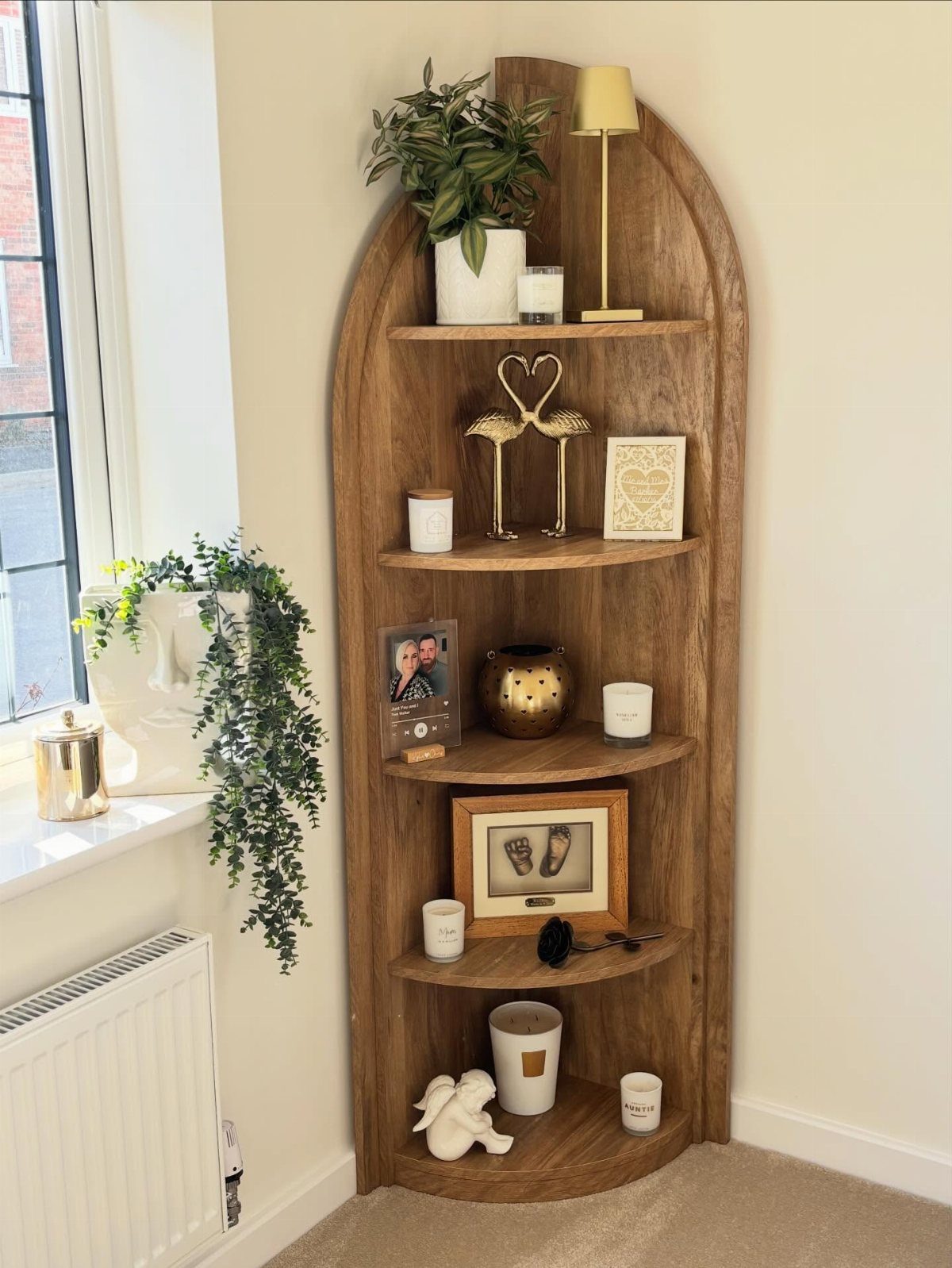 Wooden arched corner shelving unit holding decorative plants, candles, and framed photographs.