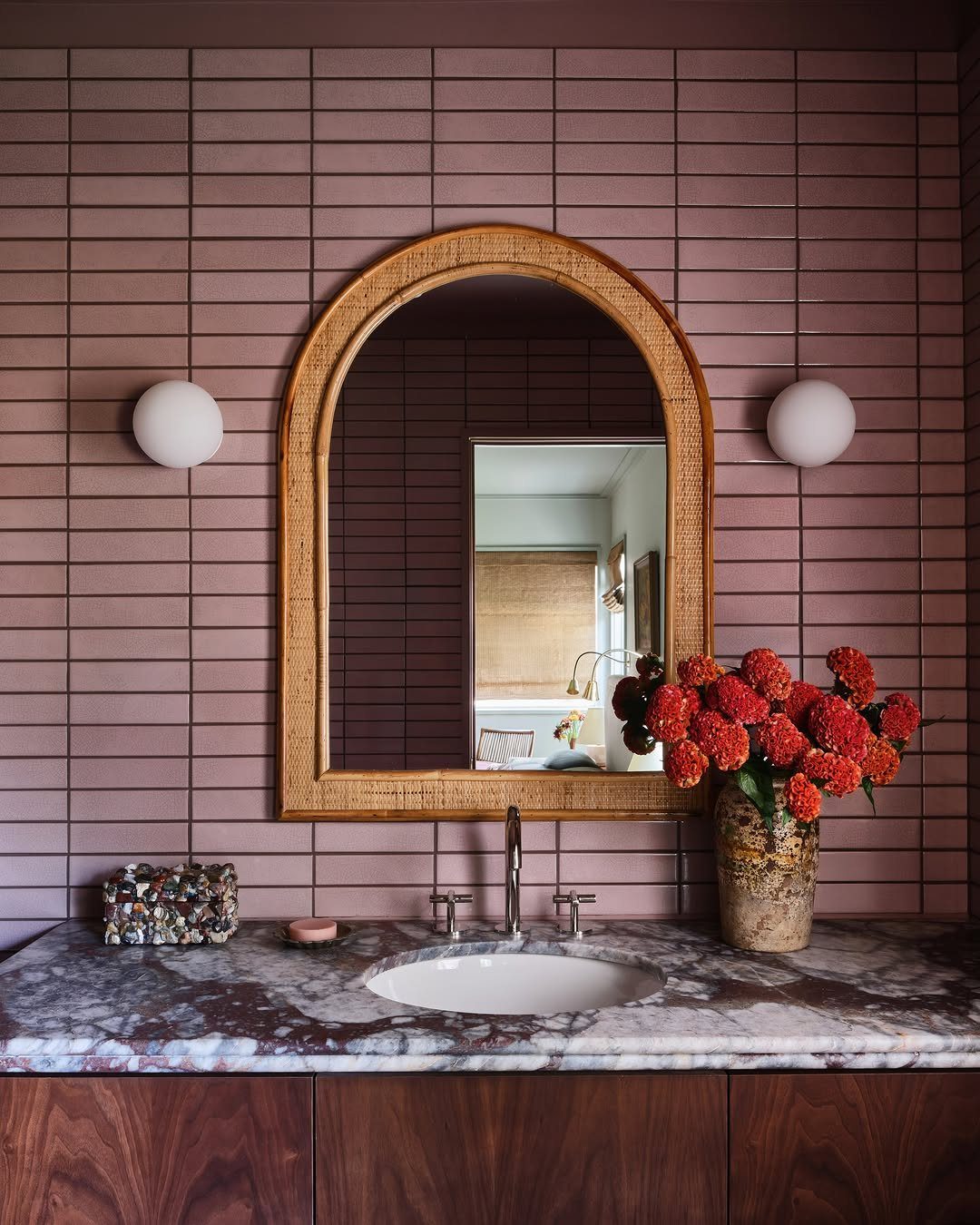 Dusty rose tiled bathroom wall with an arched rattan mirror and marble countertop.