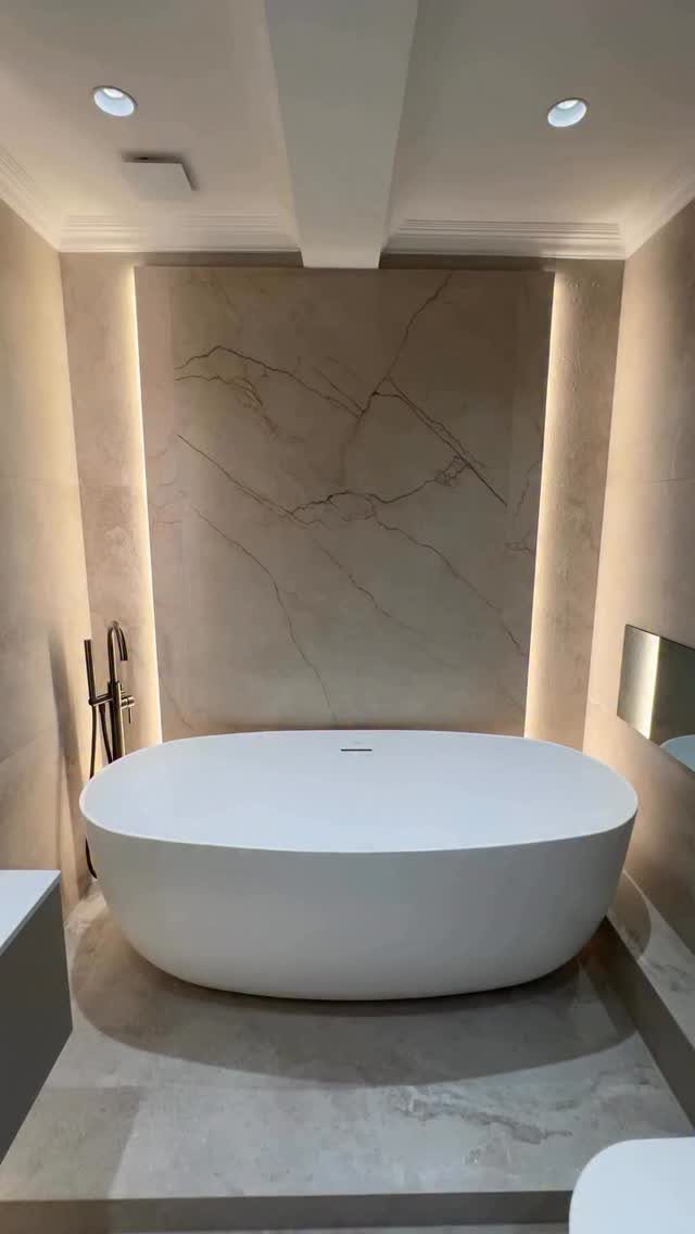 White oval freestanding bathtub against a backlit beige marble wall in a modern bathroom