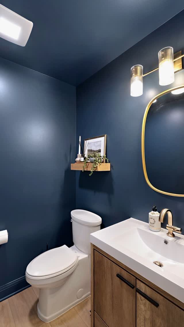 Deep blue bathroom walls with a gold-rimmed mirror and modern gold light fixture.