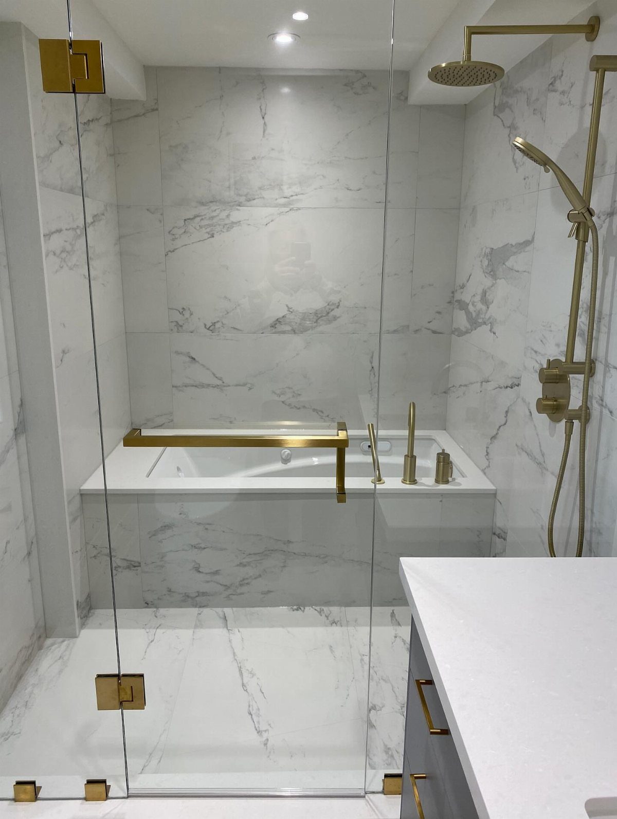 Modern bathroom with white marble tiles, gold fixtures, and a glass shower enclosure.