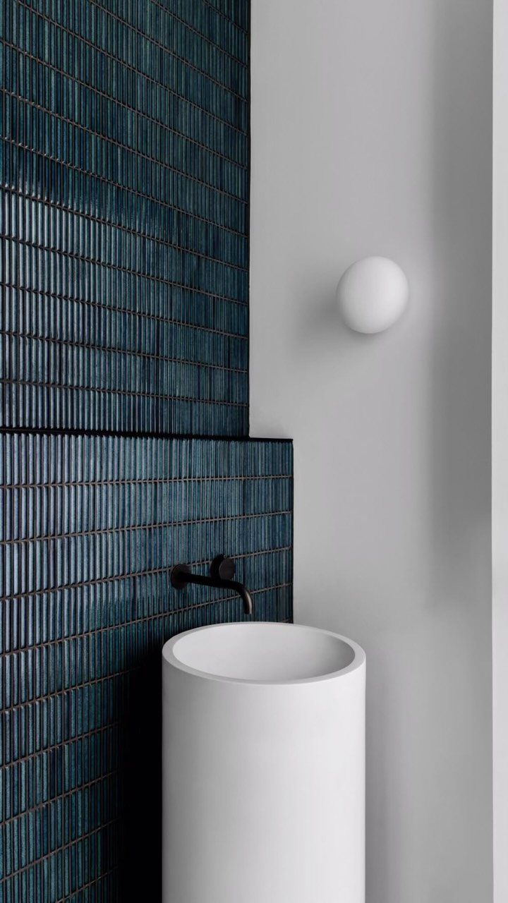 White cylindrical pedestal sink with black faucet against dark blue tiled wall