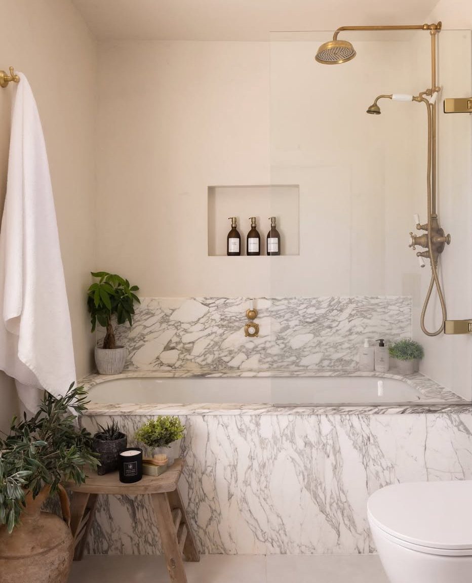 White bathtub with marble surround and gold shower fixtures in a bright bathroom.