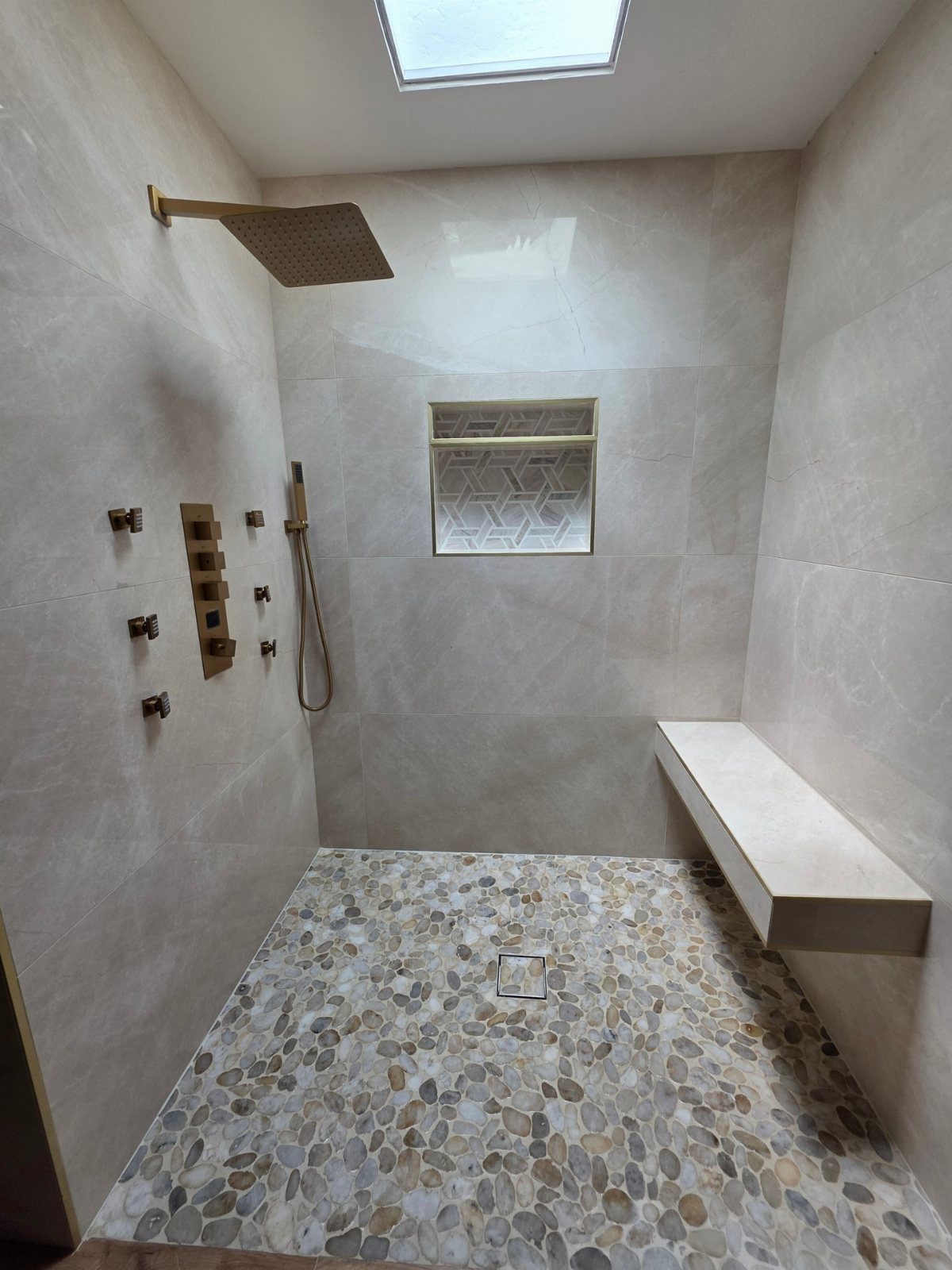 Walk-in shower featuring a pebble stone floor and beige marble walls with gold fixtures.