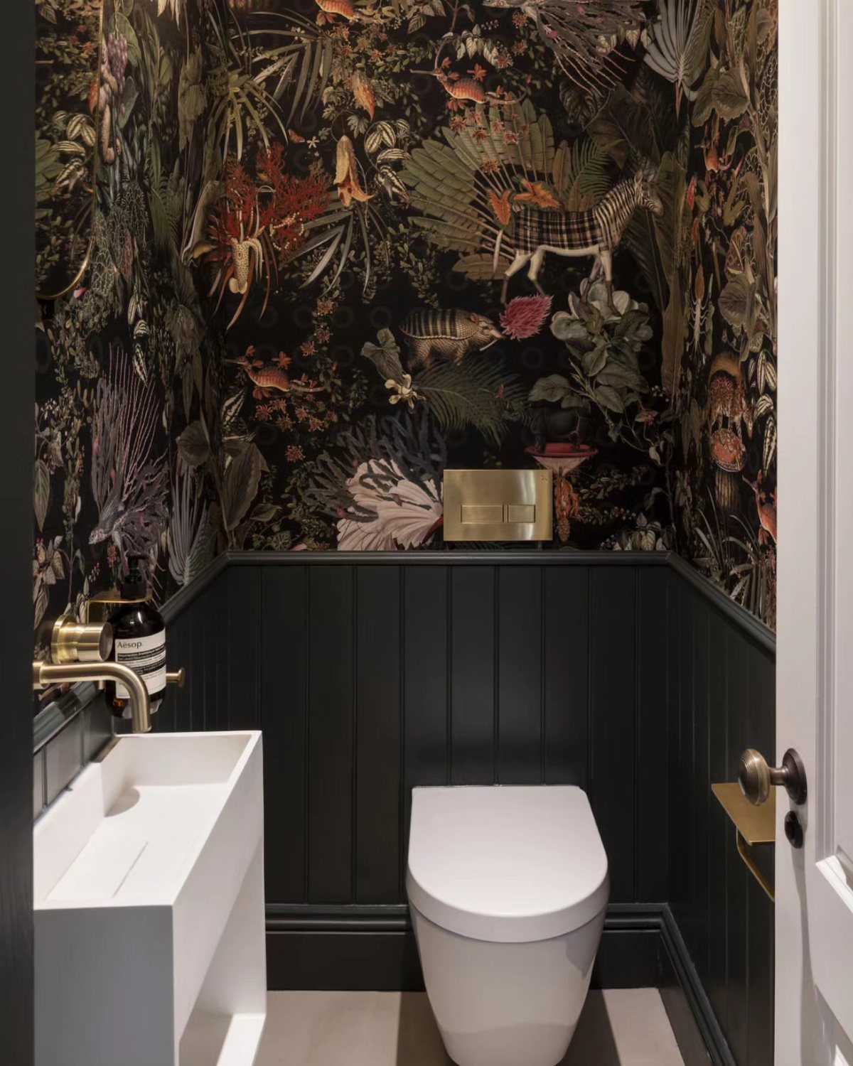 Dark jungle patterned wallpaper in a bathroom with black wainscoting and white fixtures.