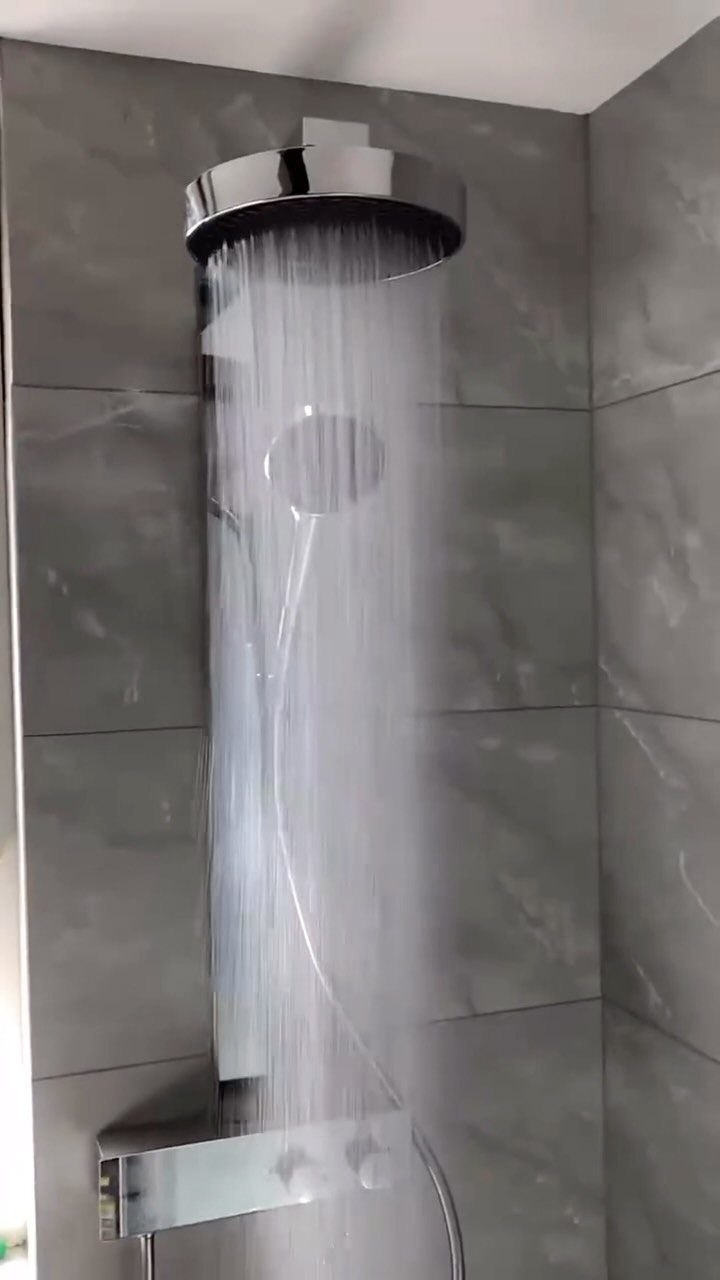 Chrome rain shower head with water streaming against grey tiled bathroom walls.