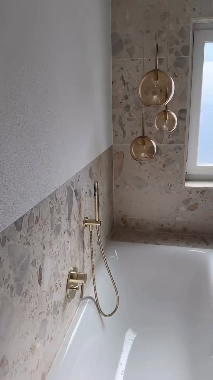 White bathtub with beige terrazzo tile walls and gold bathroom fixtures