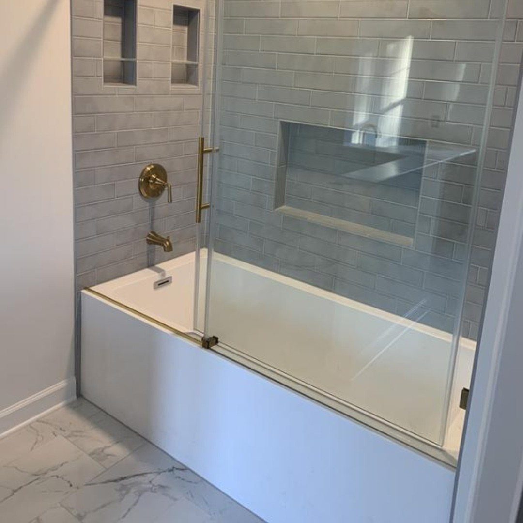 White alcove bathtub with grey tiled walls, gold fixtures, and glass door.
