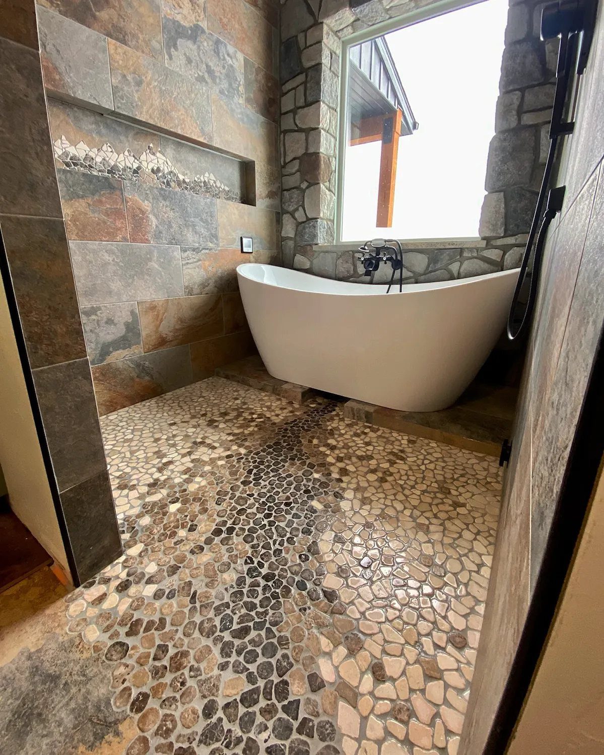 White freestanding bathtub surrounded by earthy natural stone wall tiles and pebble flooring.