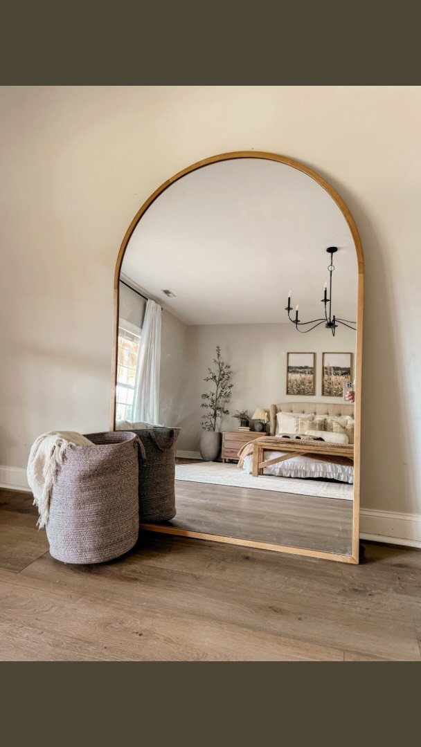 Arched gold mirror reflecting a bedroom with a bed and neutral decor.