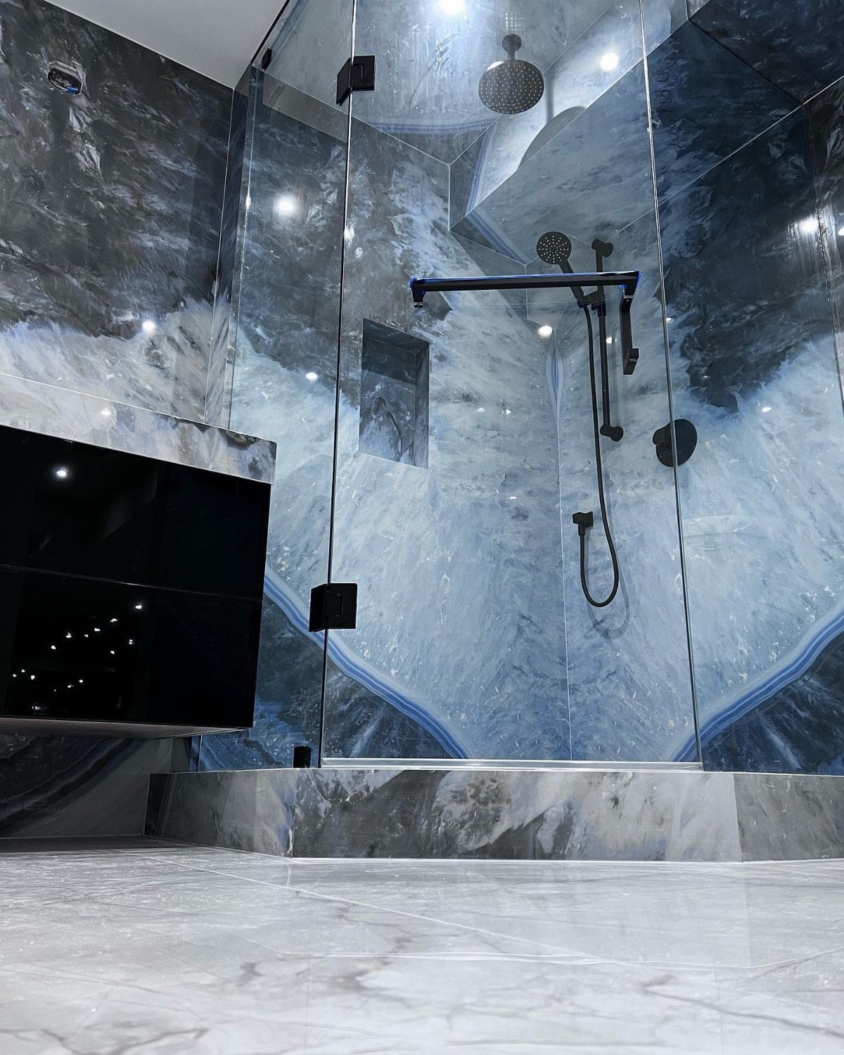 Glass shower enclosure with blue and grey marbled stone walls and white marble floors.