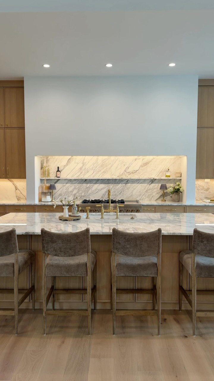 Modern kitchen with white marble surfaces, light wood cabinets, and grey bar stools.