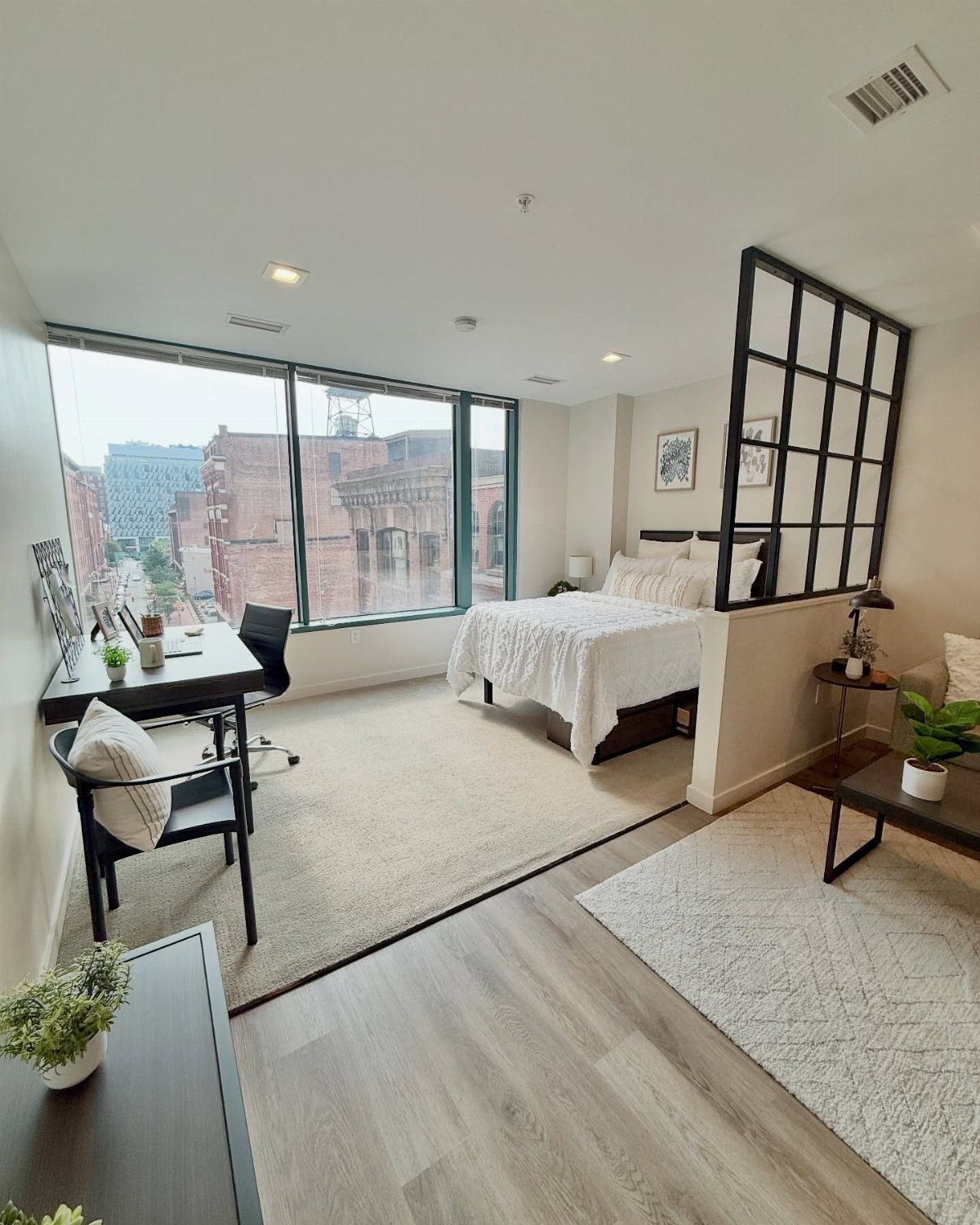 Bright studio apartment with a bed, desk, and large windows overlooking city buildings.