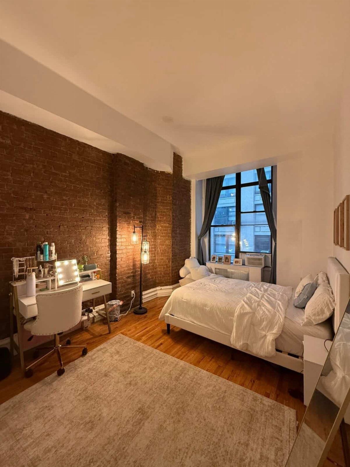 Small studio apartment with white bedding, a vanity, and an exposed brick wall.