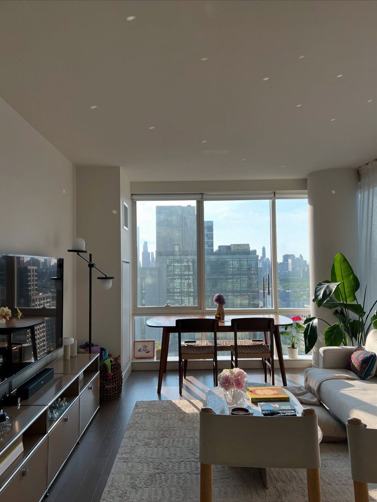 Modern apartment interior with a dining table and large window overlooking city skyscrapers.
