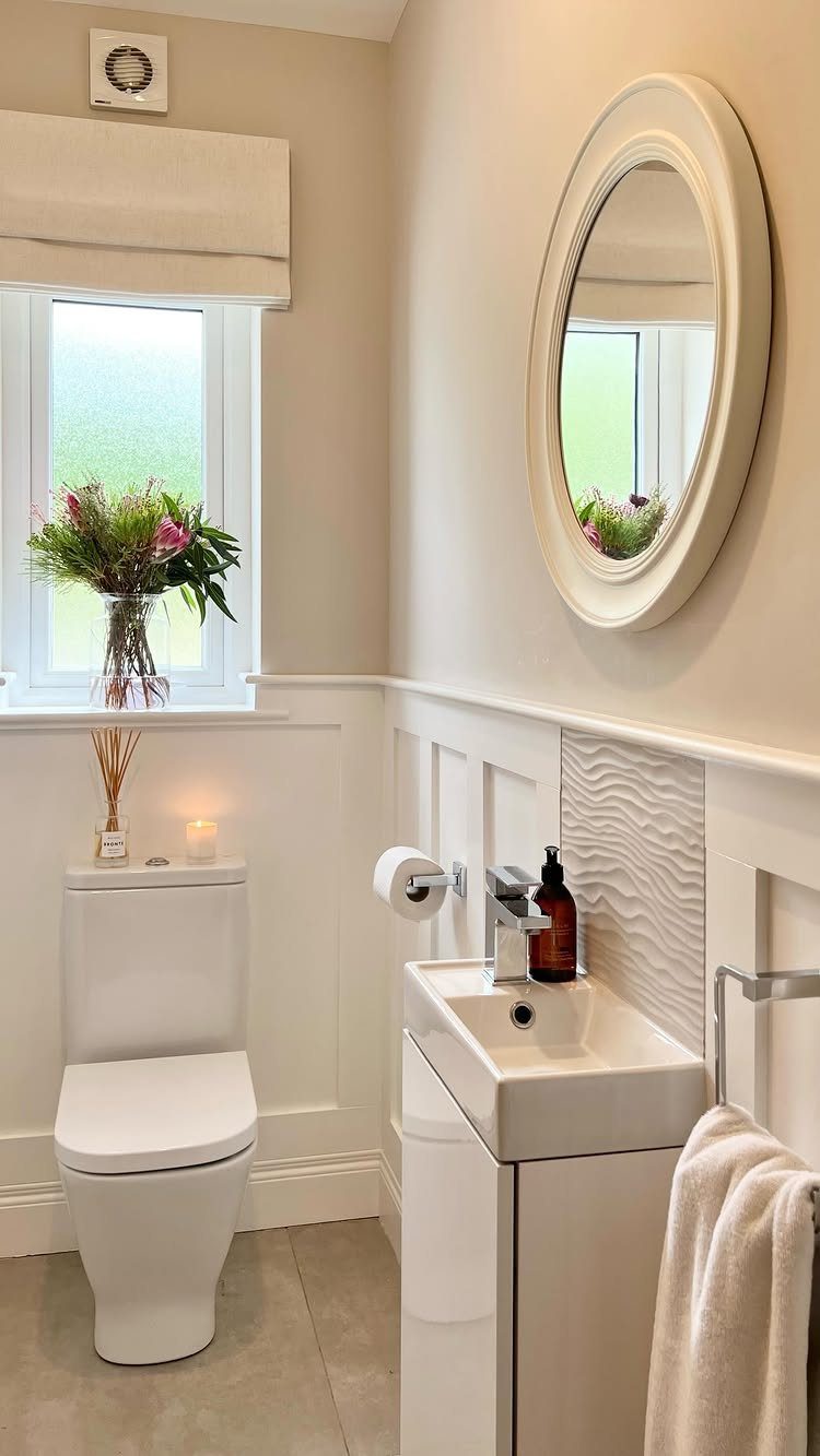 White bathroom with a vanity sink, toilet, oval mirror, and fresh flowers.