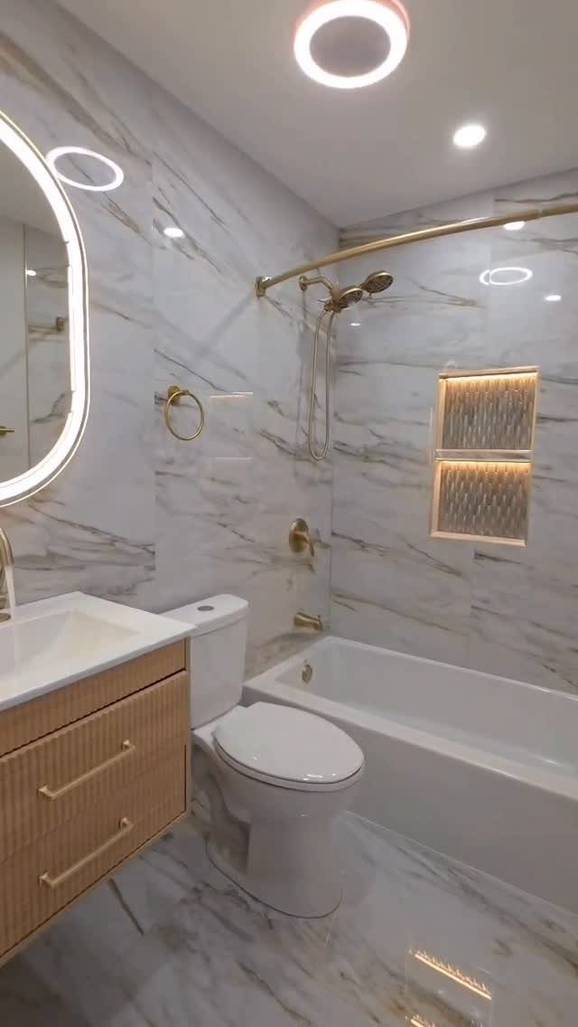 White marble bathroom with gold fixtures, a light wood vanity, and round mirror.