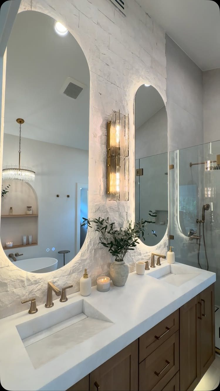 Two large oval backlit mirrors above a white double vanity with gold fixtures.