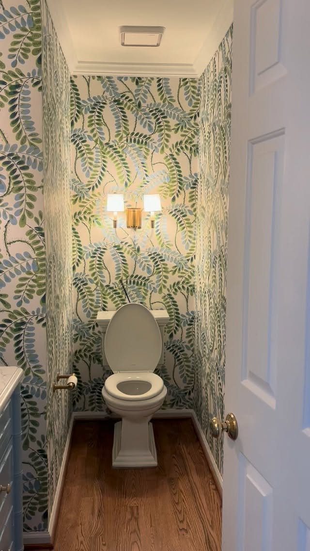 Small bathroom with blue and green botanical wallpaper, white toilet, and hardwood floors.