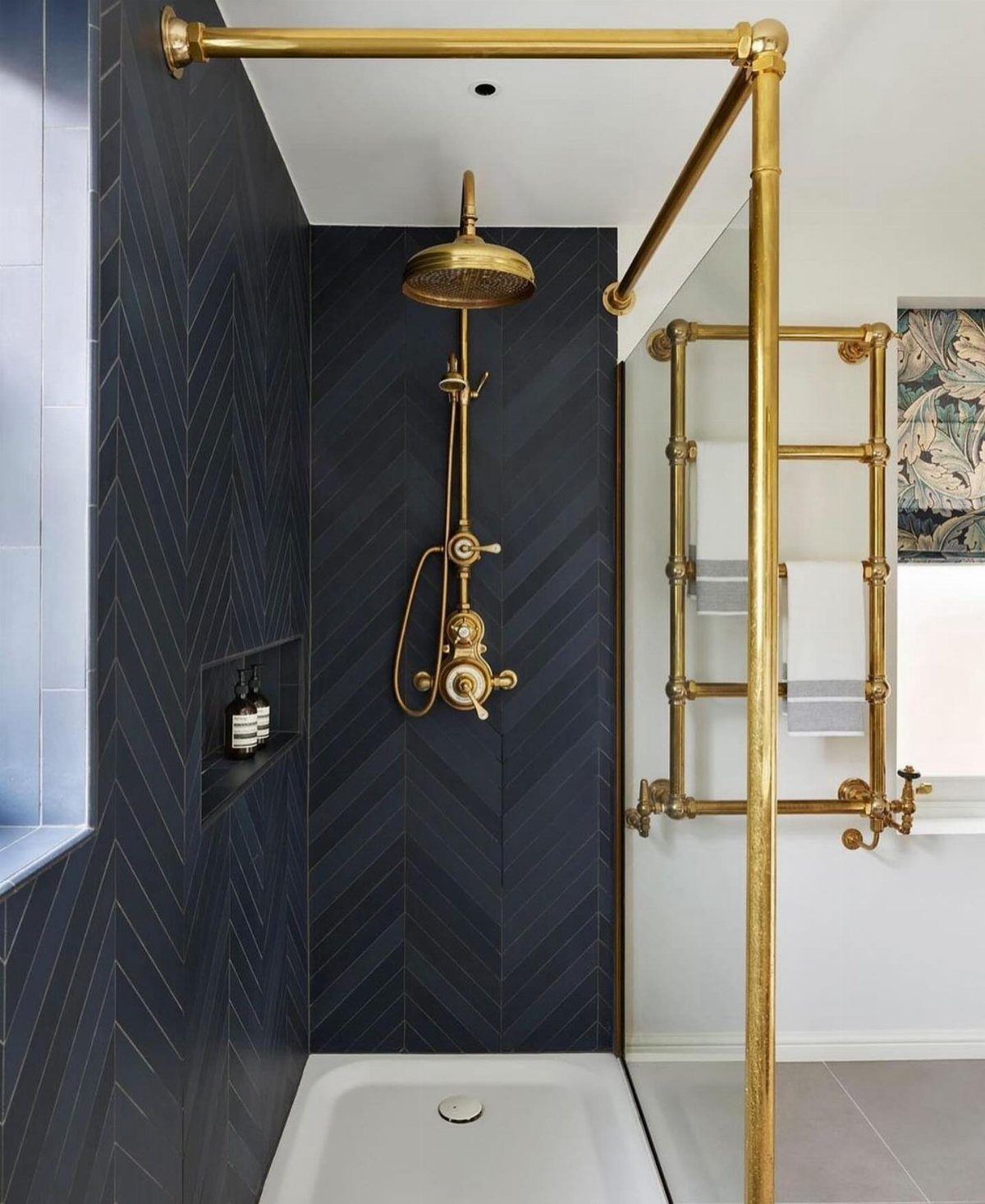 Dark blue herringbone tiled shower with gold fixtures and floral wallpaper accent.