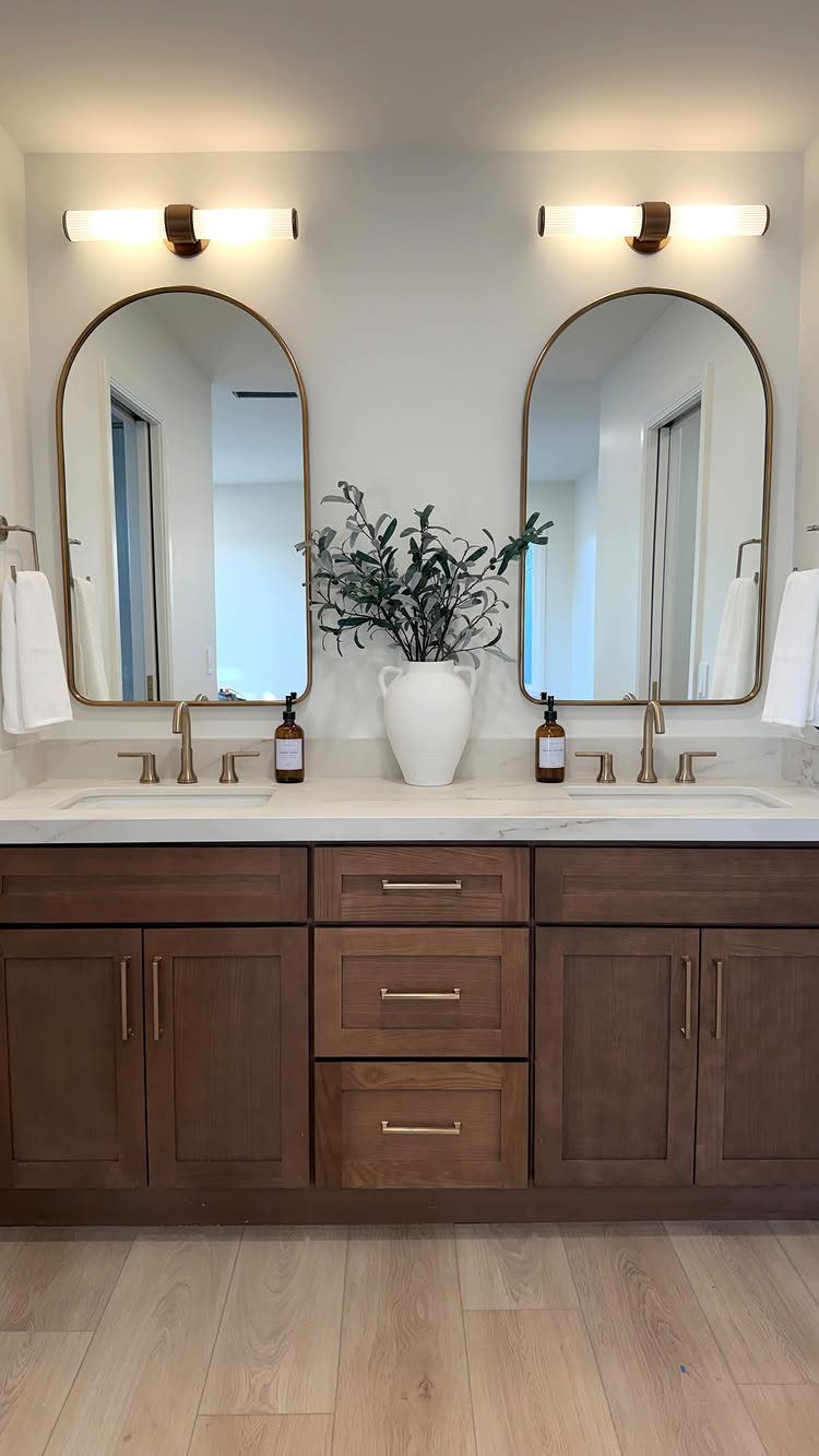 Brown wooden double sink bathroom vanity with white marble top and arched mirrors.