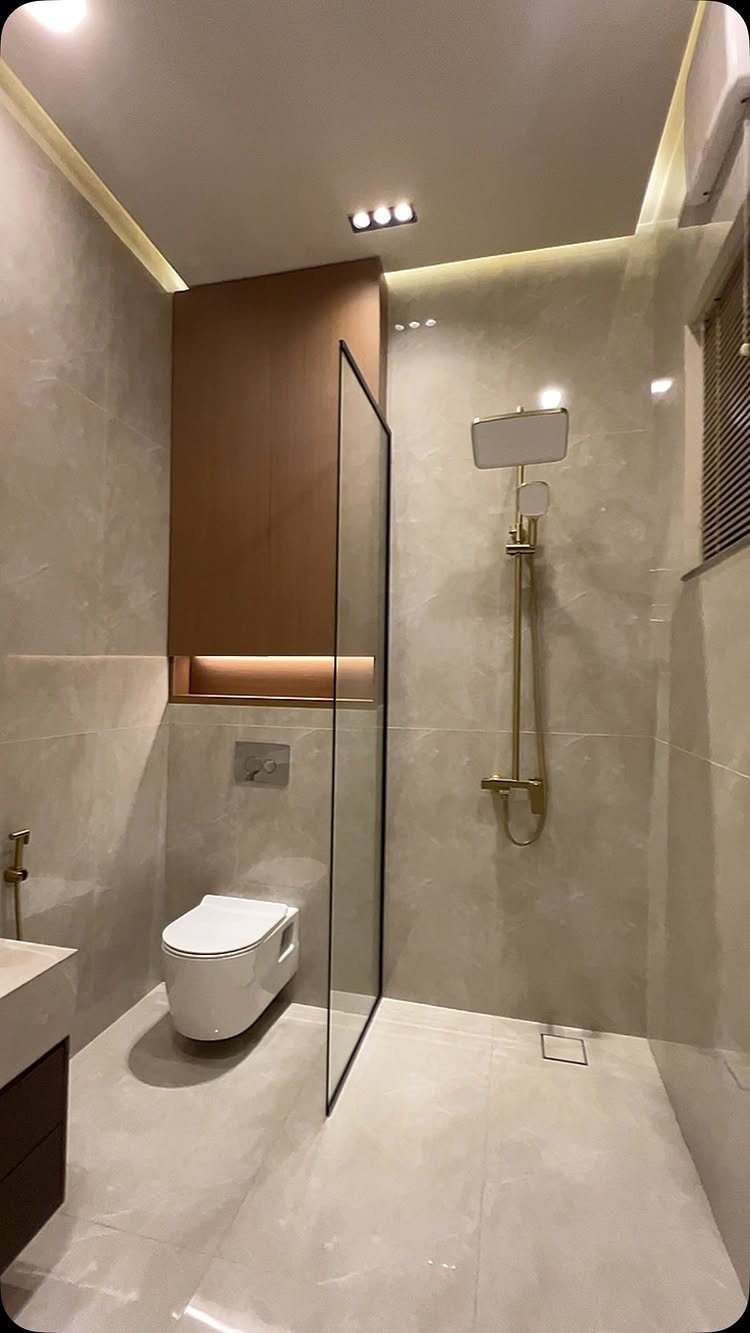 Modern bathroom with light beige polished tiles, gold fixtures, and a glass partition.