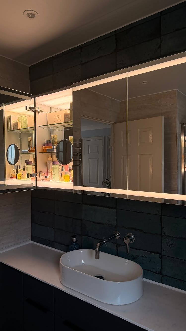 Mirrored bathroom cabinet with integrated lighting above a white vessel sink and dark tiles.