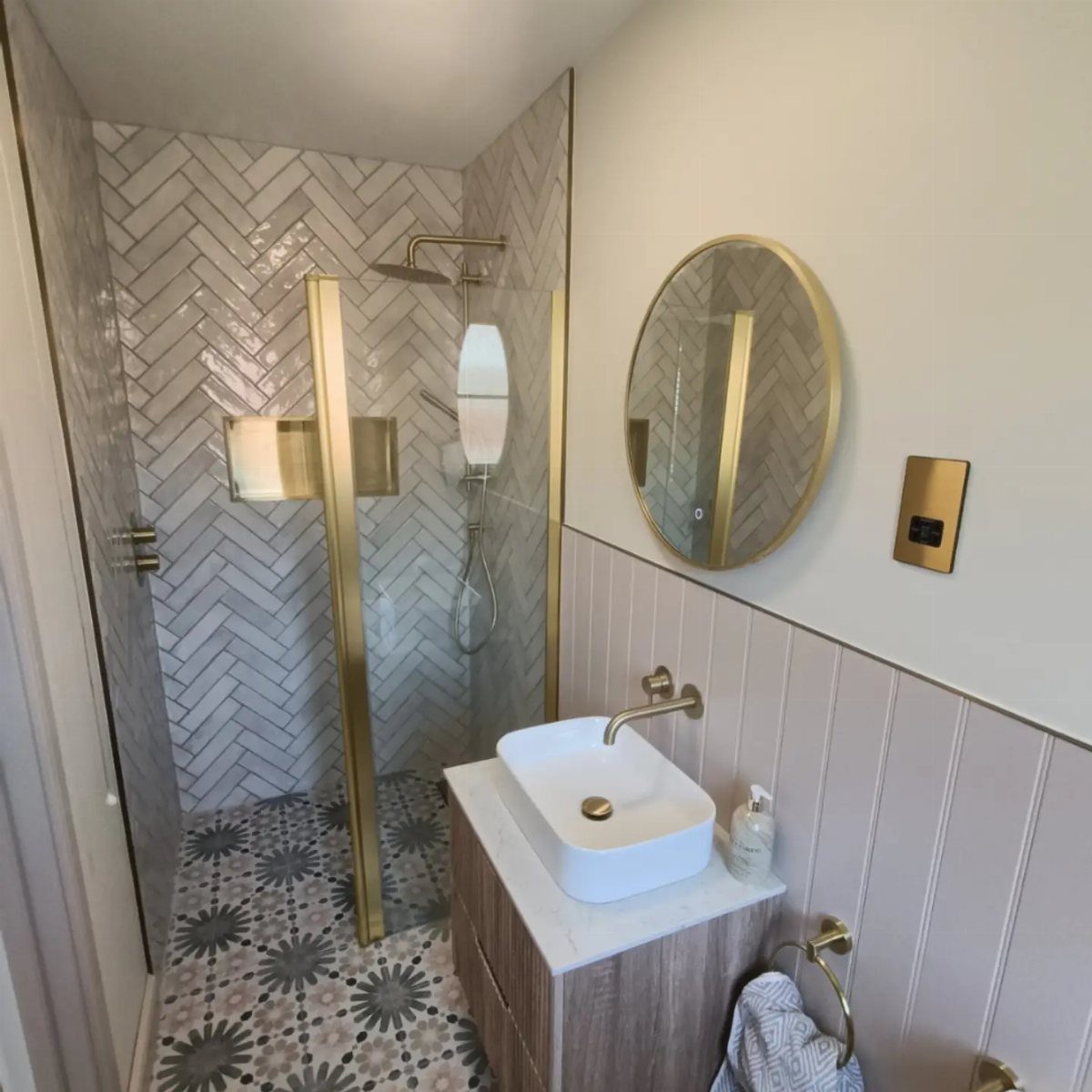 Modern bathroom with gold fixtures, herringbone wall tiles, and a glass walk-in shower.