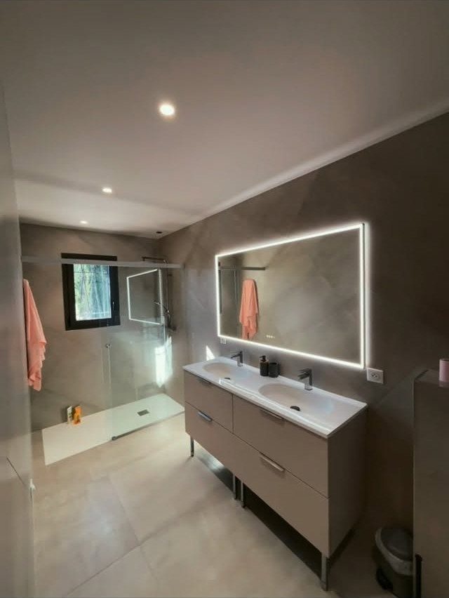Modern bathroom with large beige floor tiles, grey vanity, and backlit mirror.