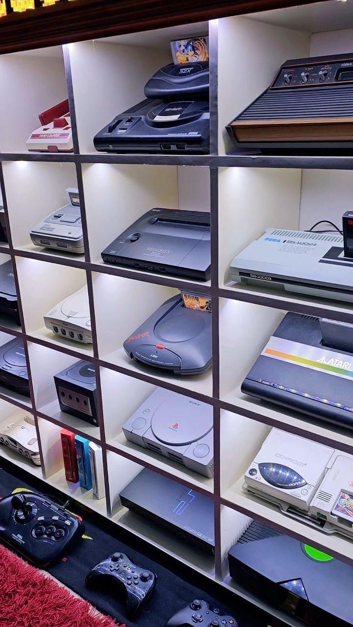 Gaming consoles and controllers displayed on a white illuminated shelving unit.