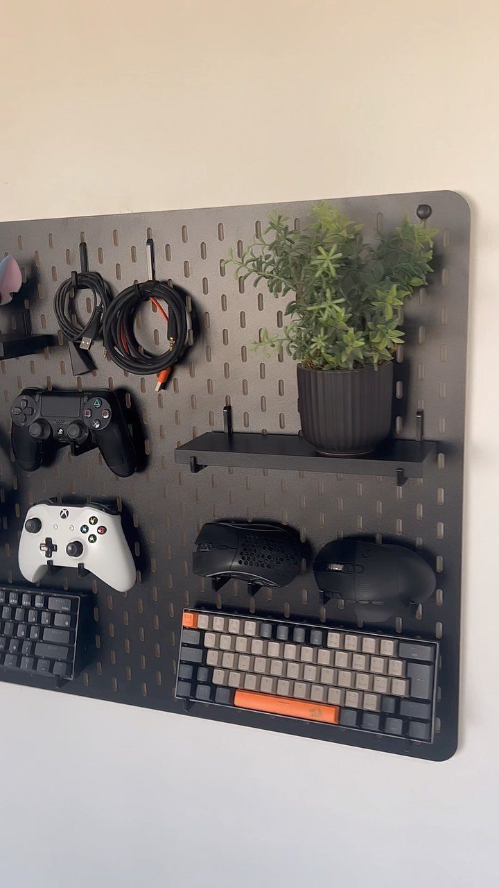 Black pegboard organizing gaming controllers, keyboard, and mouse on a white wall.