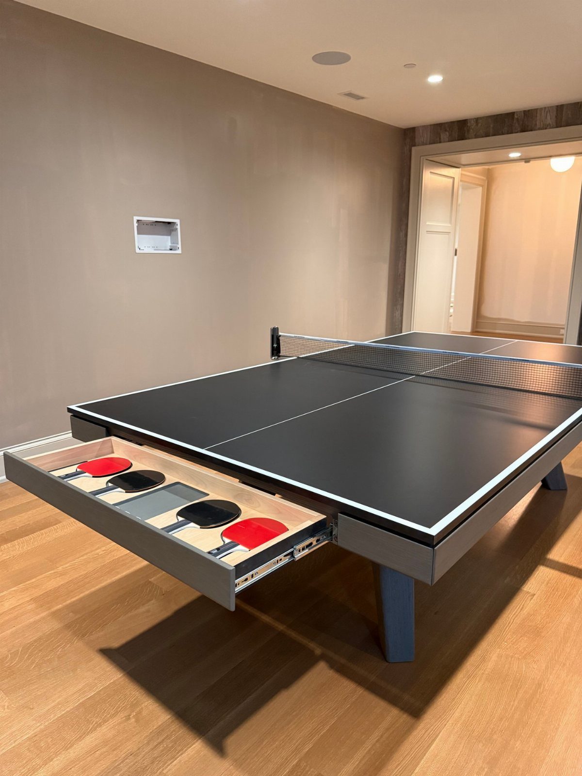 Ping pong table with a built-in storage drawer for paddles on wood flooring.