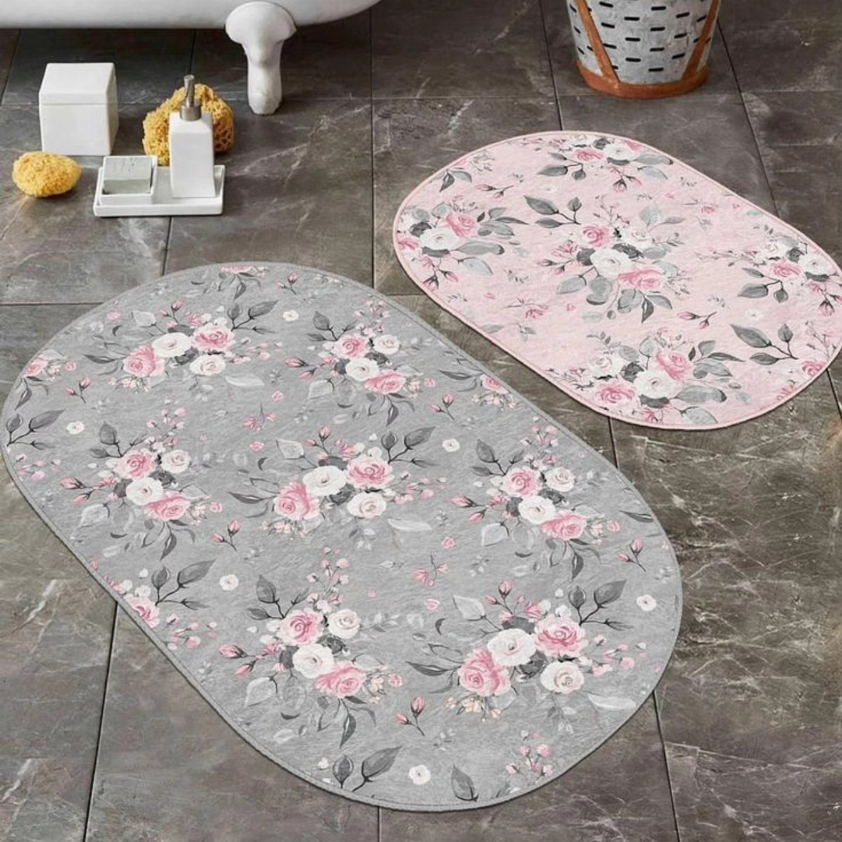 Two oval floral bath mats in grey and pink on dark tiled flooring.
