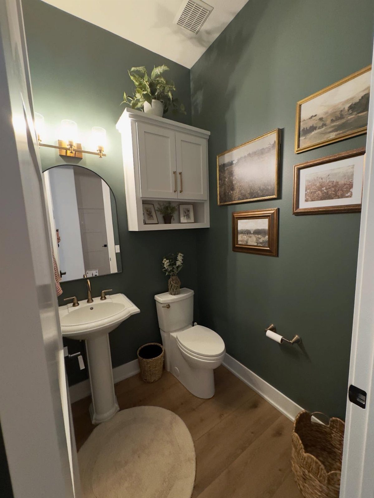 Half bathroom with dark green walls and three framed landscape paintings on the wall.