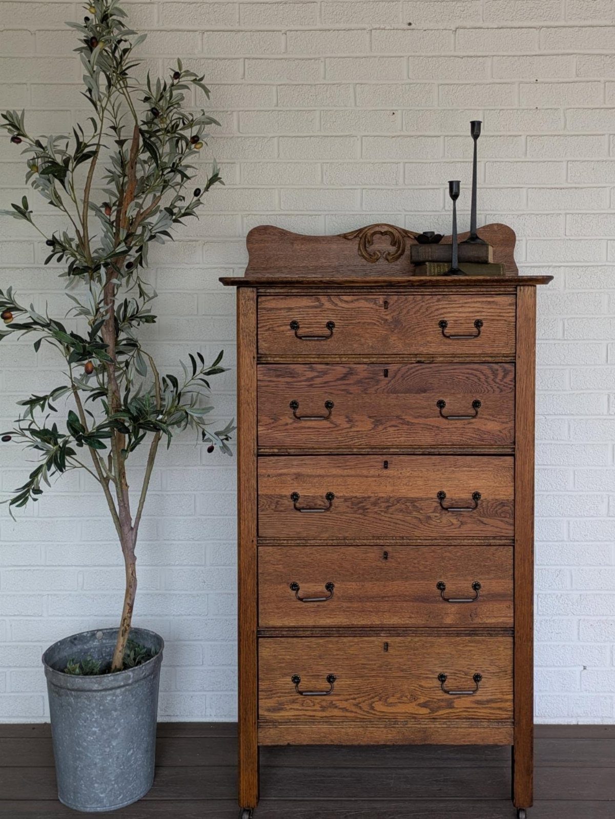 Tall wooden dresser with five drawers next to a potted olive tree.