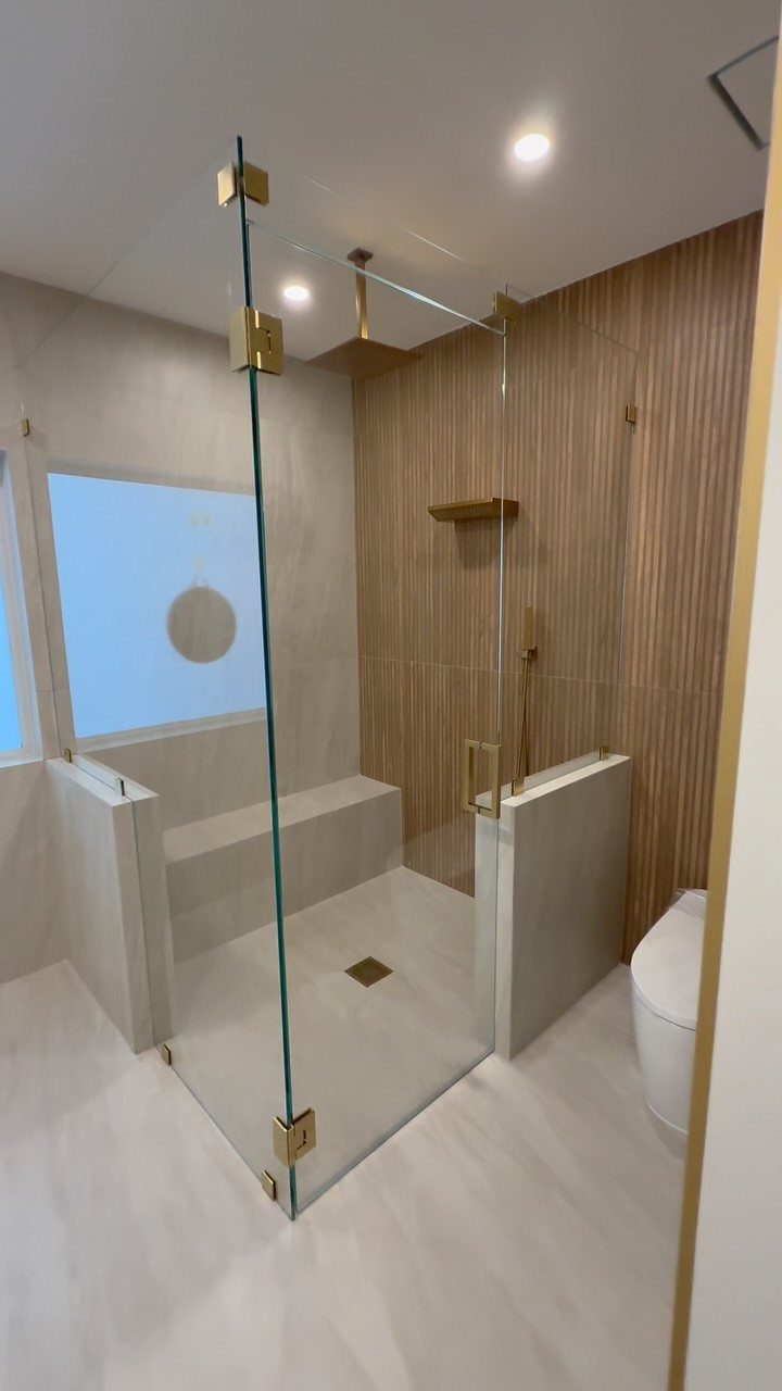 Frameless glass shower doors with gold hardware in a modern white and wood bathroom