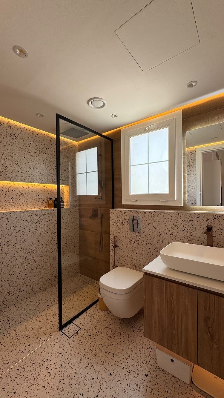 Modern bathroom with light terrazzo surfaces, wood vanity, and warm accent lighting.