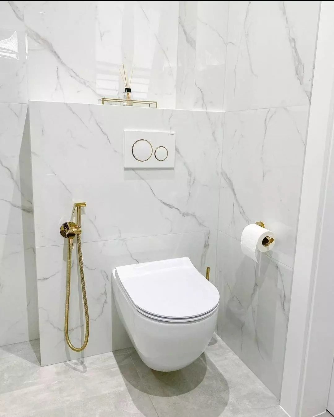 Wall-mounted white toilet with gold fixtures against white marble tiled bathroom walls.