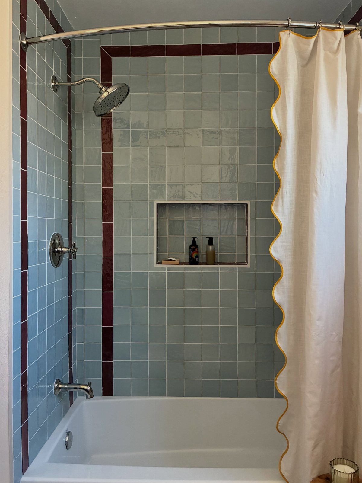 Bathtub and shower with light blue tiles and dark red accents in a bathroom.