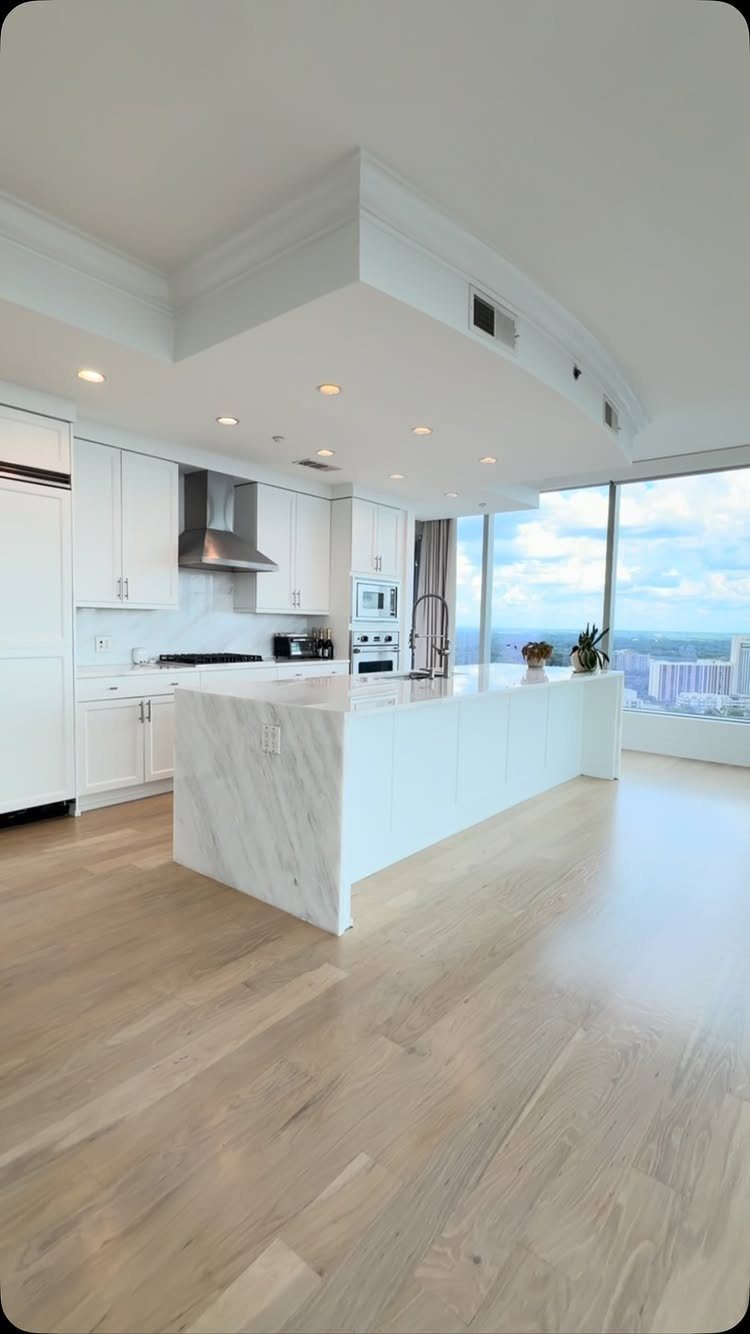 Modern white kitchen with marble island and large windows overlooking a city skyline.